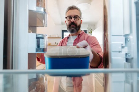 Handsome man putting lunchbox in fridge. Taking out lunch from fridge, eating leftovers. Do not trow food away, reducting food waste.