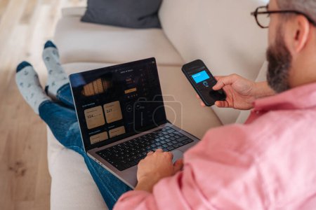 Photo for Man with notebook and smartphone sitting on sofa, online banking, internetbanking at home throught banks website and app. Paying with smartphone online, contactless payments by mobile phone, virtual - Royalty Free Image