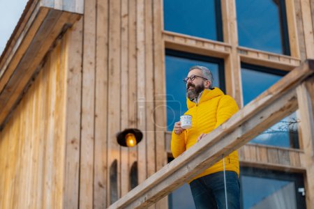 Photo for Mature man standing on cabins patio, drinking hot tea, coffee and enjoying beautiful winter day. Handsome man spending relaxing, stress-free winter weekend in cabin in mountains, enjoying alone time. - Royalty Free Image