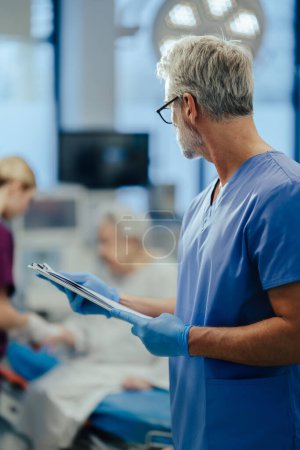 Photo for Mature male doctor looking at patient in hospital bed. ER doctor examining senior patient, reading her medical test, lab results in clipboard. ER with patient with life threatening health problems. - Royalty Free Image