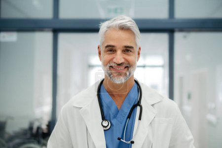 Photo for Portrait of confident mature doctor standing in Hospital corridor. Handsome doctor with gray hair wearing white coat, scrubs, stethoscope around neck standing in modern private clinic, looking at - Royalty Free Image