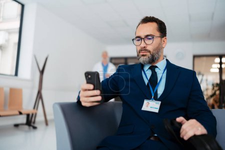 Pharmaceutical sales representative sitting in medical building, waiting for the doctor, presenting new pharmaceutical product. Male drug rep sitting in hall scrolling on smartphone.