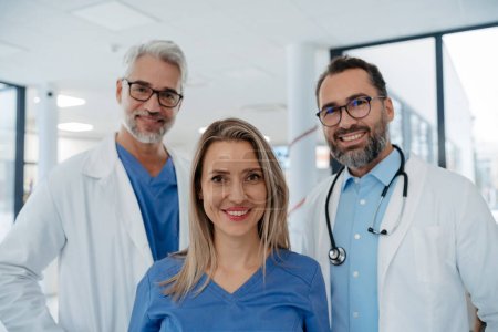 Photo for Portrait of confident three doctors standing in Hospital corridor. Medical team wearing white coat, stethoscope around neck standing in modern private clinic, looking at camera, smiling, - Royalty Free Image