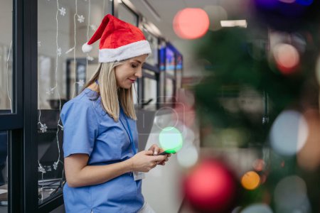 Photo for Portrait of beautiful nurse with christmas hat in hospital corridor, holding smartphone. Working Working in hospital on the Christmas day, Christmas Eve. Female doctor working a Christmas shift and - Royalty Free Image