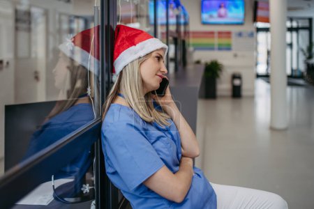 Photo for Portrait of beautiful nurse with christmas hat making call. Working in hospital on the Christmas day, Christmas Eve. Female doctor working a Christmas shift and cant be with her family. - Royalty Free Image