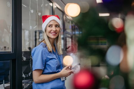 Téléchargez les photos : Portrait de belle infirmière avec chapeau de Noël dans le couloir de l'hôpital, tenant smartphone. Travailler à l'hôpital le jour de Noël, la veille de Noël. Médecin de sexe féminin travaillant un quart de Noël et - en image libre de droit