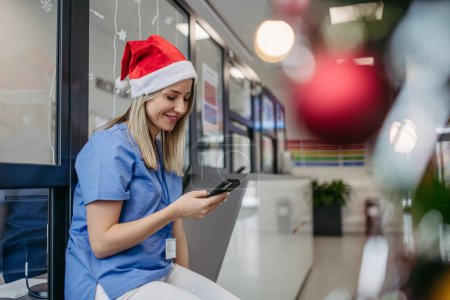 Téléchargez les photos : Portrait de belle infirmière avec chapeau de Noël dans le couloir de l'hôpital, tenant smartphone, lecture de SMS. Travailler à l'hôpital le jour de Noël, la veille de Noël. Médecin féminin travaillant un - en image libre de droit