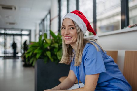 Foto de Hermosa enfermera con sombrero de Navidad sentada en la sala de espera del hospital, pasillo. Enfermera obstétrica trabajando en el hospital el día de Navidad, Nochebuena. Mujer médico trabajando un turno de Navidad y puede - Imagen libre de derechos
