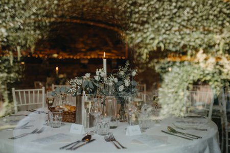 Foto de Elegante y romántico entorno de mesa para la recepción de bodas interiores con luces de hadas en la pared de ladrillo detrás. Decoración de mesa de boda de lujo, mesa de eventos especiales con arreglos de flores frescas. - Imagen libre de derechos