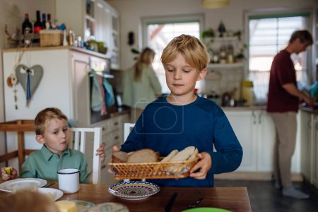Foto de Familia preparando el desayuno en la cocina casera por la mañana. Hijo mayor trayendo pan sobre la mesa. - Imagen libre de derechos