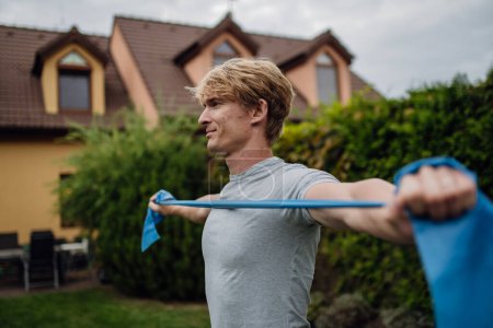 Photo for Middle aged man doing strength exercises with resistance band outdoors in the garden. Concept of workout routine at home. - Royalty Free Image