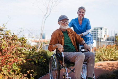 Photo for Nurse pushing senior man in a wheelchair. Female caregiver and elderly man enjoying a warm autumn day in nursing home, residential care home. - Royalty Free Image