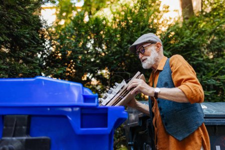 Foto de Hombre mayor arrojando residuos de papel y cartón en el contenedor de reciclaje en el apartamento delantero. Anciano clasificando los residuos de acuerdo con el material en contenedores de colores. - Imagen libre de derechos