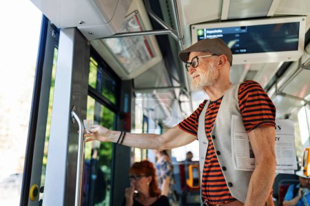 Téléchargez les photos : Homme âgé voyageant à travers la ville en bus, debout à la porte. Tramway pour se rendre à l'épicerie en utilisant les transports en commun. - en image libre de droit