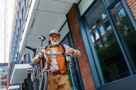 Téléchargez les photos : Un homme âgé, cycliste voyageant à travers la ville en vélo, portant le vélo sur l'épaule. Aîné banlieue en vélo. - en image libre de droit