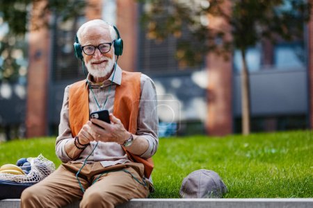 Téléchargez les photos : Portrait d'un homme âgé élégant assis dans le parc de la ville, écoutant de la musique via un casque. Concept de vieil homme jeune de cœur. - en image libre de droit