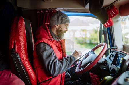 Téléchargez les photos : Chauffeur de camion assis dans le camion, tenant la tablette, regardant les détails de la cargaison, calendrier de livraison. Concept de gestion de charge. - en image libre de droit