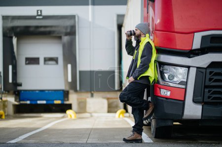 Téléchargez les photos : Chauffeur de camion appuyé contre son camion rouge et boire du café, attendant les travailleurs d'entrepôt. - en image libre de droit