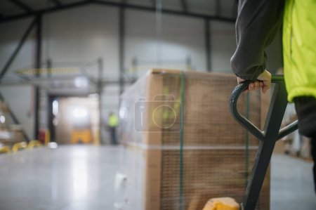 Téléchargez les photos : Gros plan d'un ouvrier d'entrepôt manœuvrant un cric à palettes chargé de marchandises à travers l'entrepôt. - en image libre de droit