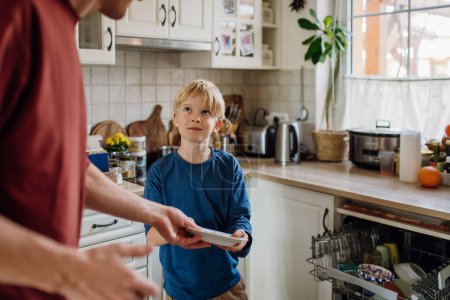 Téléchargez les photos : Jeune fils aidant père à charger le lave-vaisselle après le petit déjeuner. Nettoyage de la cuisine avant de partir au travail et à l'école. routine matinale familiale. - en image libre de droit