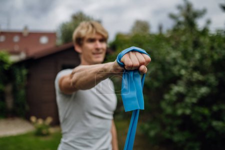 Foto de Hombre de mediana edad haciendo ejercicios de fuerza con banda de resistencia al aire libre en el jardín. Concepto de rutina de entrenamiento en casa. - Imagen libre de derechos