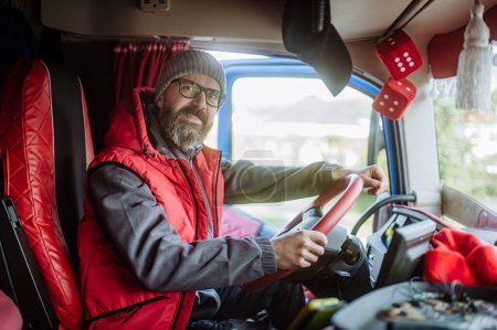 Téléchargez les photos : Portrait d'un camionneur professionnel confiant assis dans une cabine de camion, regardant la caméra et souriant. À l'intérieur du véhicule. - en image libre de droit