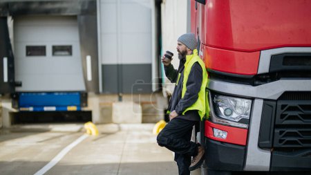 Foto de Conductor de camión apoyado en su camión rojo y beber café, esperando a los trabajadores del almacén. - Imagen libre de derechos