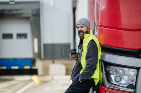 Téléchargez les photos : Chauffeur de camion appuyé contre son camion rouge et boire du café, attendant les travailleurs d'entrepôt. - en image libre de droit