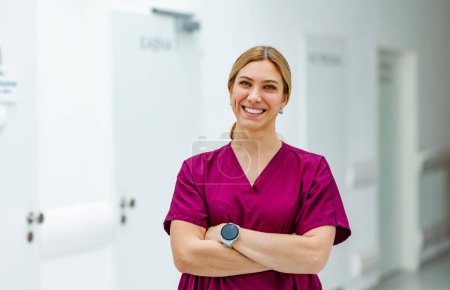 Téléchargez les photos : Portrait d'une belle infirmière en uniforme, debout dans le couloir de l'hôpital les bras croisés, regardant la caméra et souriant. - en image libre de droit