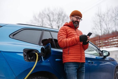 Reifer Mann lädt Elektroauto an kalten, verschneiten Tagen, mit einer Ladeapp für Elektrofahrzeuge, überprüft den Energieverbrauch, die Akkulaufzeit auf dem Smartphone. Laden und Fahren von Elektrofahrzeugen während
