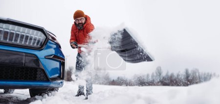Téléchargez les photos : Un homme libérant sa voiture coincée dans la neige avec une pelle, sortant la voiture de la dérive de neige. Ramasser et jeter de la neige, pelleter. Bannière d'hiver avec espace de copie. - en image libre de droit