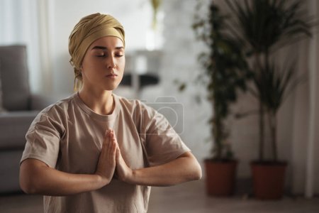 Foto de Mujer joven con cáncer tomando yoga y meditando en su apartamento. Paciente femenina fuerte calmando su mente con ejercicio fácil. Concepto de salud mental y cáncer. Día de concienciación. - Imagen libre de derechos