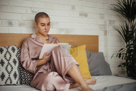 Photo for Young woman with cancer reading book. Concept of cancer awareness. Female cancer patient lying in bed, relaxing at home. - Royalty Free Image