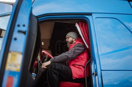 Photo for Portrait of confident professional truck driver sitting in a truck cabin, looking at camera and smiling. - Royalty Free Image