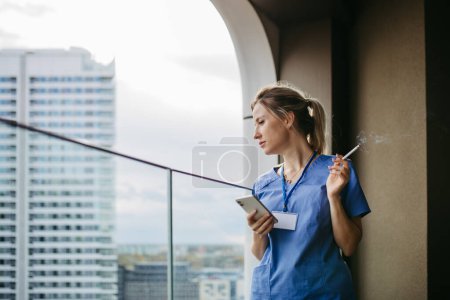 Foto de Enfermera fumando cigarrillo en el balcón después del trabajo. El médico se siente abrumado y agotado en el trabajo, tomando un descanso y fumando. - Imagen libre de derechos