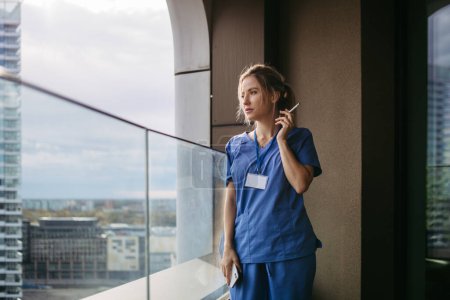 Photo for Female nurse smoking cigarette at balcony after work. Doctor feeling overwhelmed and exhausted at work, taking break and smoking. - Royalty Free Image