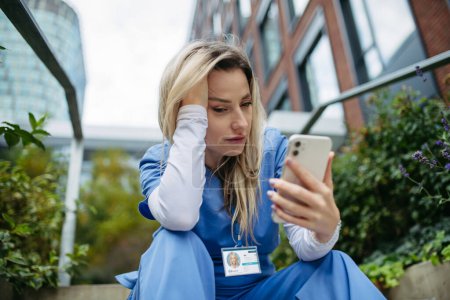 Photo for Young female doctor feeling overwhelmed at work, sitting on concrete stairs, looking at phone. Healthcare workers having stressful job, feeling exhausted. Burnout syndrome for doctors and nurses. - Royalty Free Image