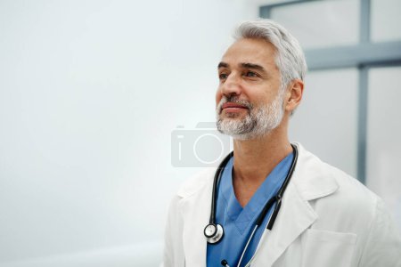 Photo for Portrait of confident mature doctor standing in Hospital corridor. Handsome doctor with gray hair wearing white coat, scrubs, stethoscope around neck standing in modern private clinic, looking at - Royalty Free Image