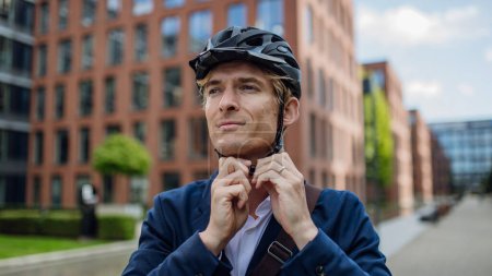 Téléchargez les photos : Un bel homme d'âge moyen se déplaçant à vélo dans la ville, attachant un casque sur la tête. Navetteur urbain masculin voyageant du travail à vélo après une longue journée de travail. - en image libre de droit