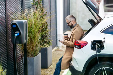 Photo for Portrait of businessman waiting while electric car is charging, sitting in car trunk, scrolling on his smartphone, and drinking coffee. An electric vehicle charging station at home. - Royalty Free Image