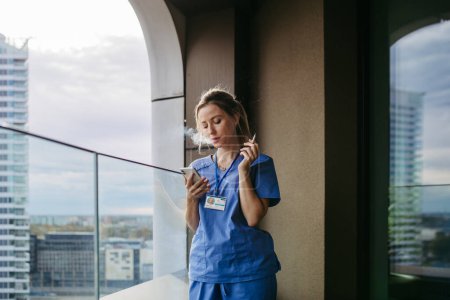 Foto de Enfermera fumando cigarrillo en el balcón después del trabajo. El médico se siente abrumado y agotado en el trabajo, tomando un descanso y fumando. - Imagen libre de derechos
