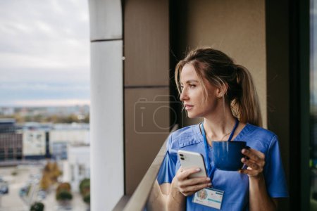 Photo for Female nurse enjoying cup of coffee at home after work, standing by window. Morning tea before work. Work-life balance for healthcare worker. Woman enjoying view from balcony. - Royalty Free Image