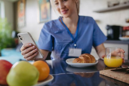 Photo for Female nurse or doctor getting ready for work in the morning, scrolling on smartphone while eating breakfast, before leaving for the work dressed in scrubs. Work-life balance for healthcare worker. - Royalty Free Image