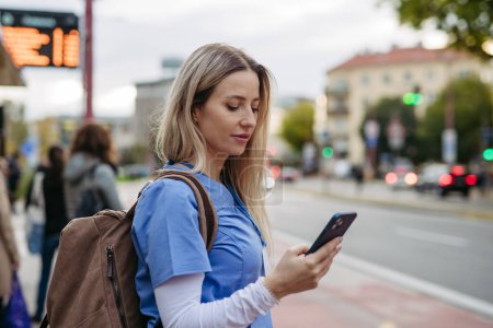 Photo for Female nurse, doctor going home from work, waiting for bus, scrolling on smartphone. Work-life balance of healthcare worker. - Royalty Free Image