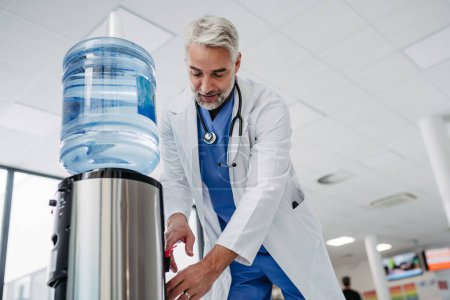 Photo for Handsome doctor is taking a break during work shift at hospital, drinking water from water dispenser in hospital lobby. - Royalty Free Image