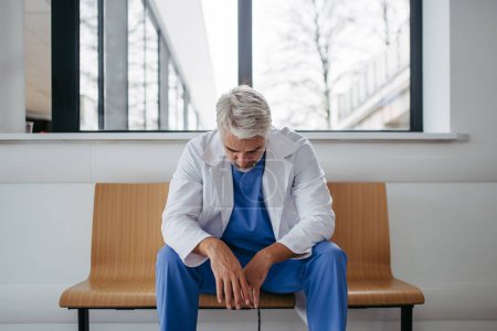 Frustrierter, erschöpfter Arzt sitzt auf Bank im Krankenhausflur. Konzept des Burnout-Syndroms bei Ärzten, Gesundheitspersonal.