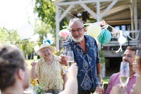 Téléchargez les photos : Grand-père claquant avec un verre de champagne. Homme âgé debout, portant un toast à la fête de jardin d'été en plein air. - en image libre de droit