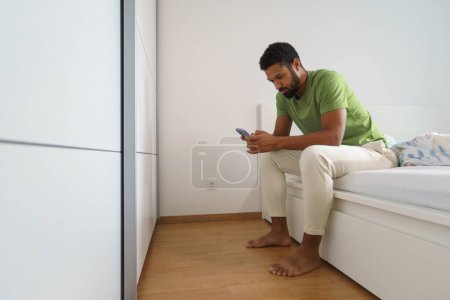 Foto de Hombre soltero despertando solo en su cama. Sentado en la cama y mirando el teléfono inteligente, leyendo mensajes de texto. - Imagen libre de derechos
