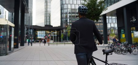 Photo for Rear view of city bike commuter leaving work on bicycle after long work day. Banner of city with copy space. - Royalty Free Image