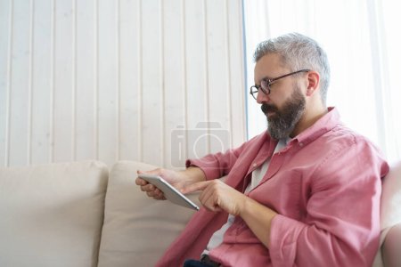 Photo for Man using smart thermostat, adjusting, lowering heating temperature at home. Man watching tv show on tablet, video streaming services with movies and tv shows online. - Royalty Free Image